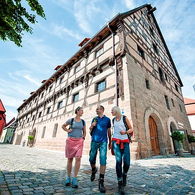 Drei Personen mit Rucksäcken gehen auf Kopfsteinpflaster vor einem Fachwerkhaus bei blauem Himmel spazieren.