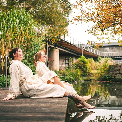 Zwei Frauen in Bademänteln sitzen am Holzsteg und halten ihre Füße ins Wasser eines Teichs im herbstlichen Park.