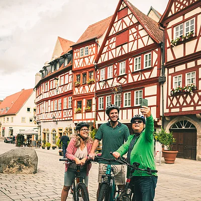 Drei Personen mit Fahrrädern machen ein Selfie vor Fachwerkhäusern in einer gepflasterten Altstadtstraße.