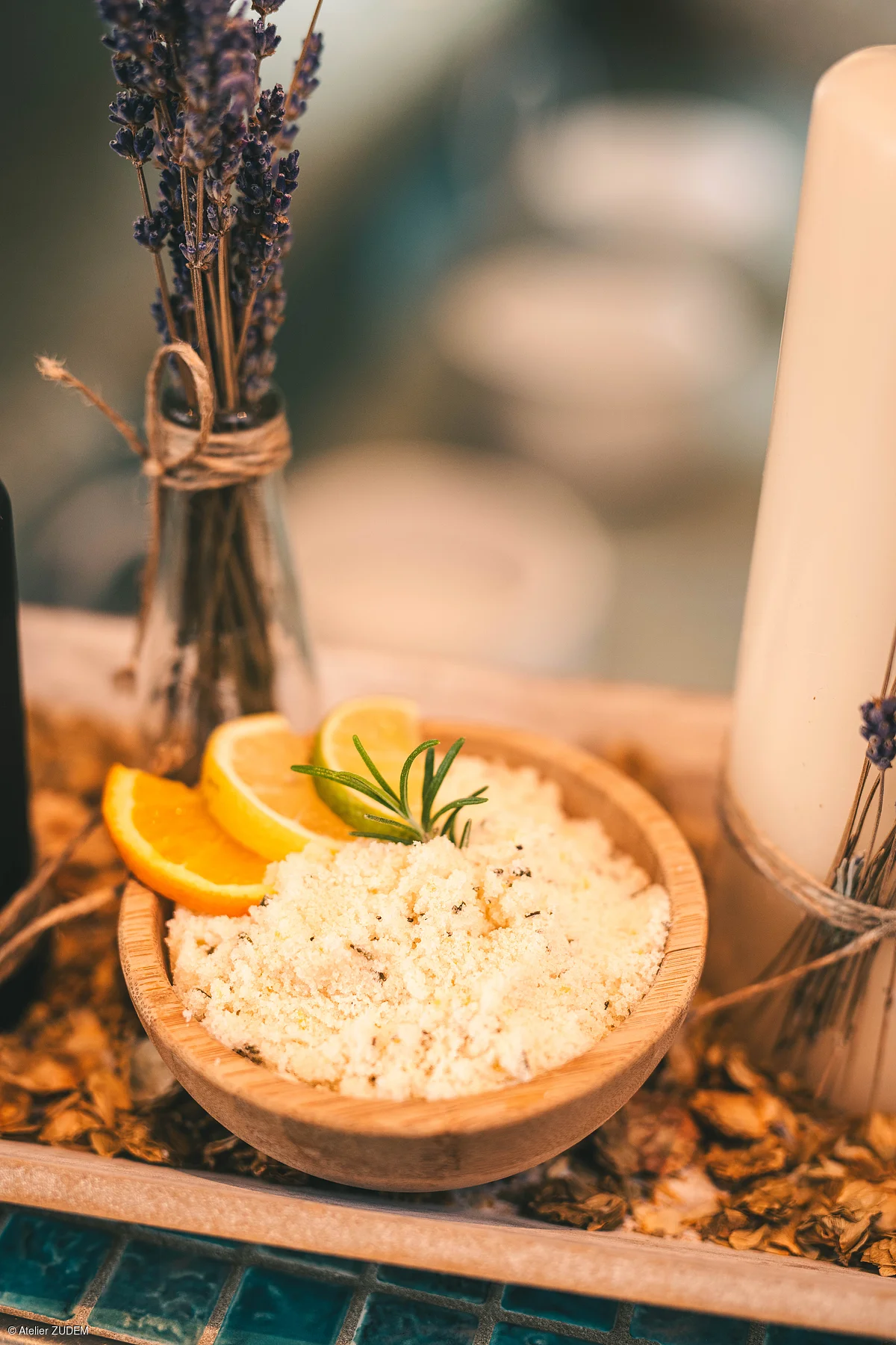 Holzschale mit Badesalz, Zitronen- und Orangenscheiben, daneben getrockneter Lavendel in Glasflasche und Kerze.