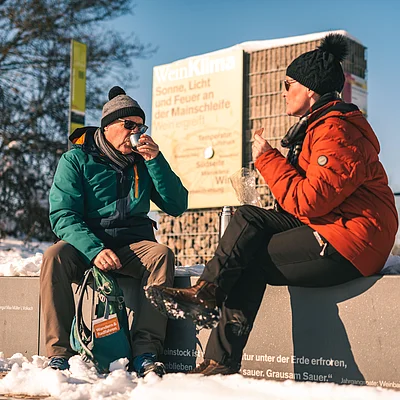 Zwei Personen in Winterkleidung sitzen auf einer Mauer im Schnee und trinken aus Bechern.