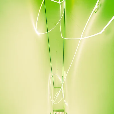 Frau geht eine beleuchtete Treppe in einem grünen, schmalen Raum mit weißen Neonlichtern an der Decke hinunter.