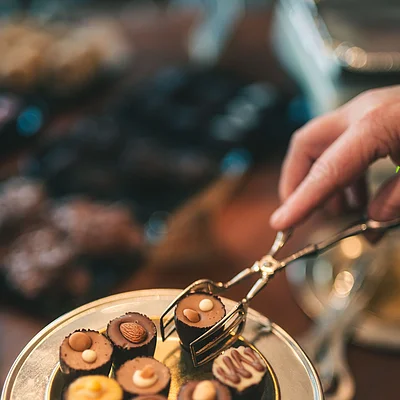 Hand mit Zange nimmt Praline von goldfarbenem Teller mit verschiedenen Pralinen darauf