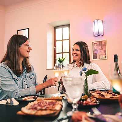 Zwei Frauen stoßen mit Weißweingläsern an einem gedeckten Tisch mit Essen und Getränken an.