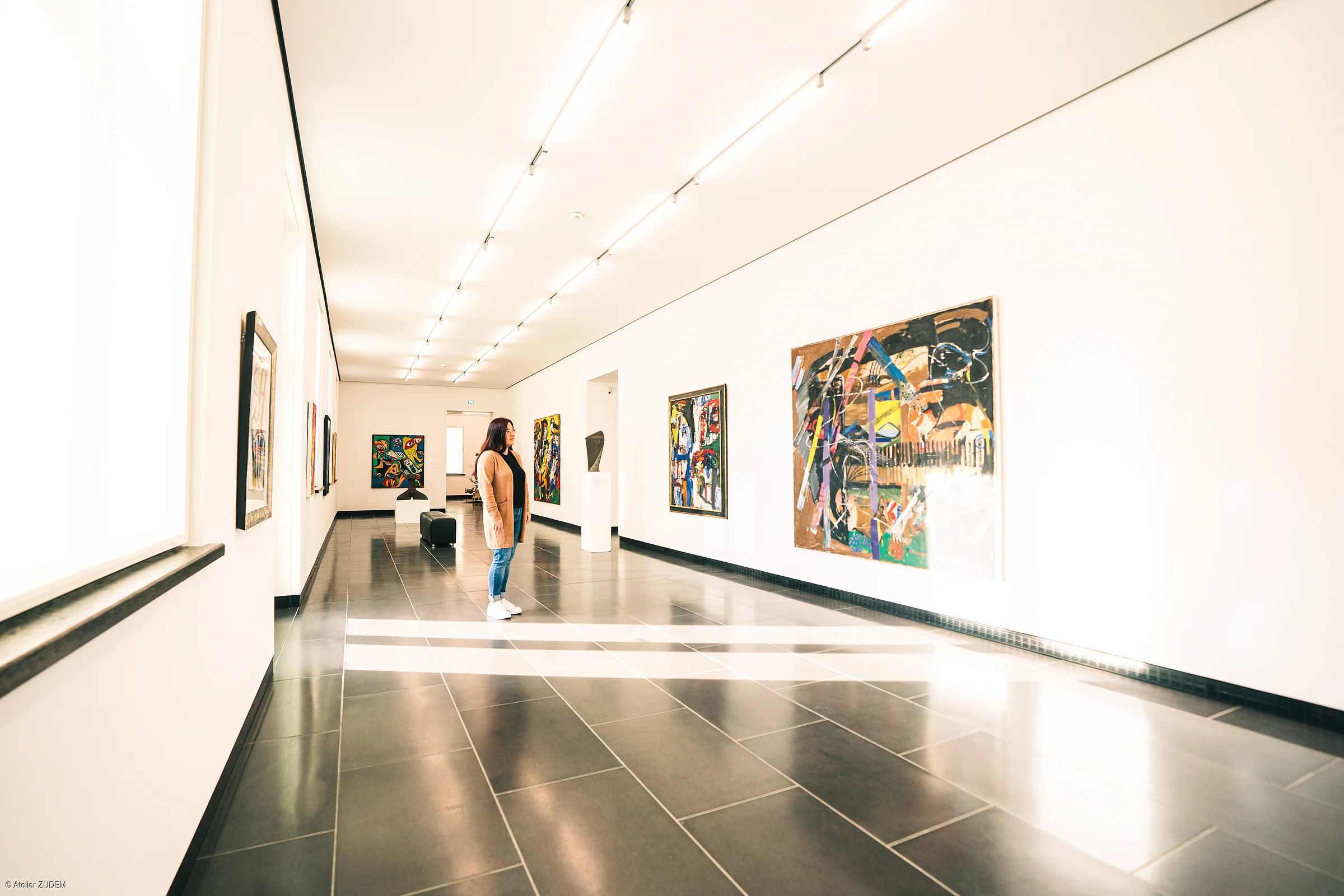 Frau betrachtet abstrakte Gemälde in einem hellen, modernen Kunstmuseum mit schwarzen Bodenfliesen.