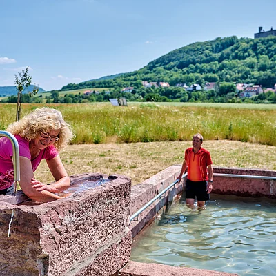 Frau wäscht Arme an Brunnen, Mann steht im Wasserbecken, Wiese und bewaldeter Hügel mit Burg im Hintergrund.