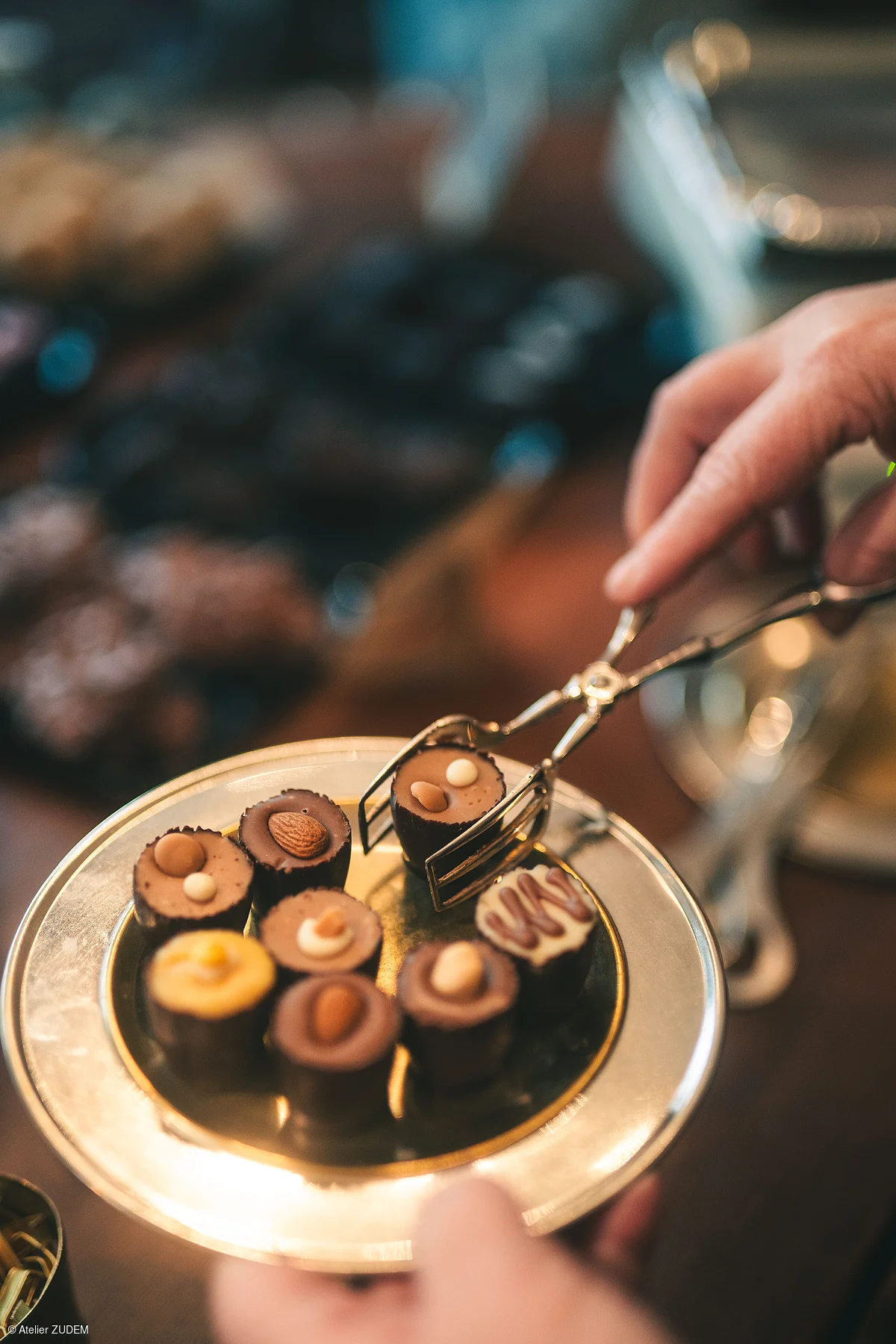 Hand mit Zange nimmt Praline von goldfarbenem Teller mit verschiedenen Pralinen darauf