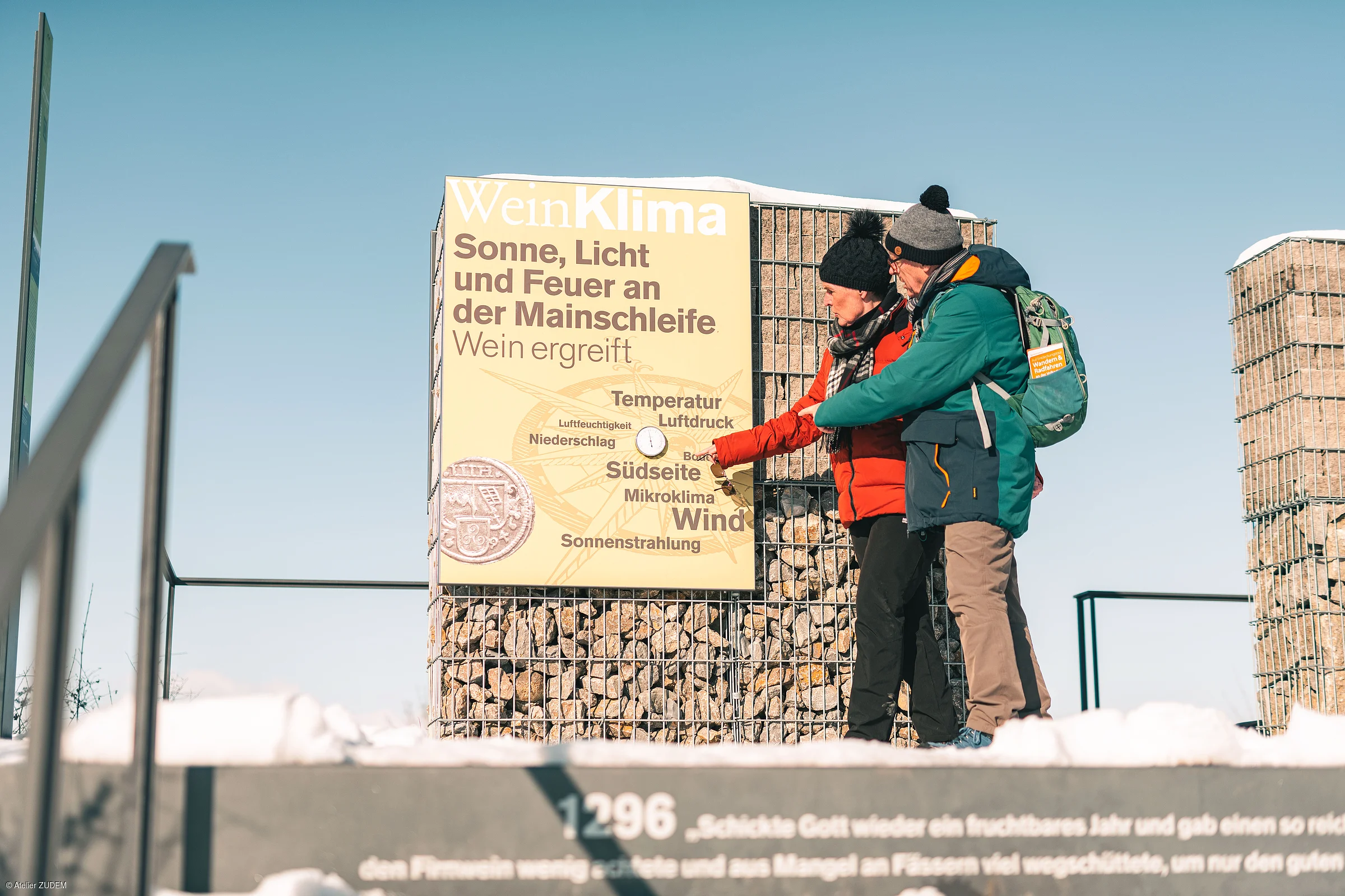Zwei Personen lesen eine Infotafel zum Thema Wein und Klima an einer steinernen Säule im Freien bei klarem Himmel.