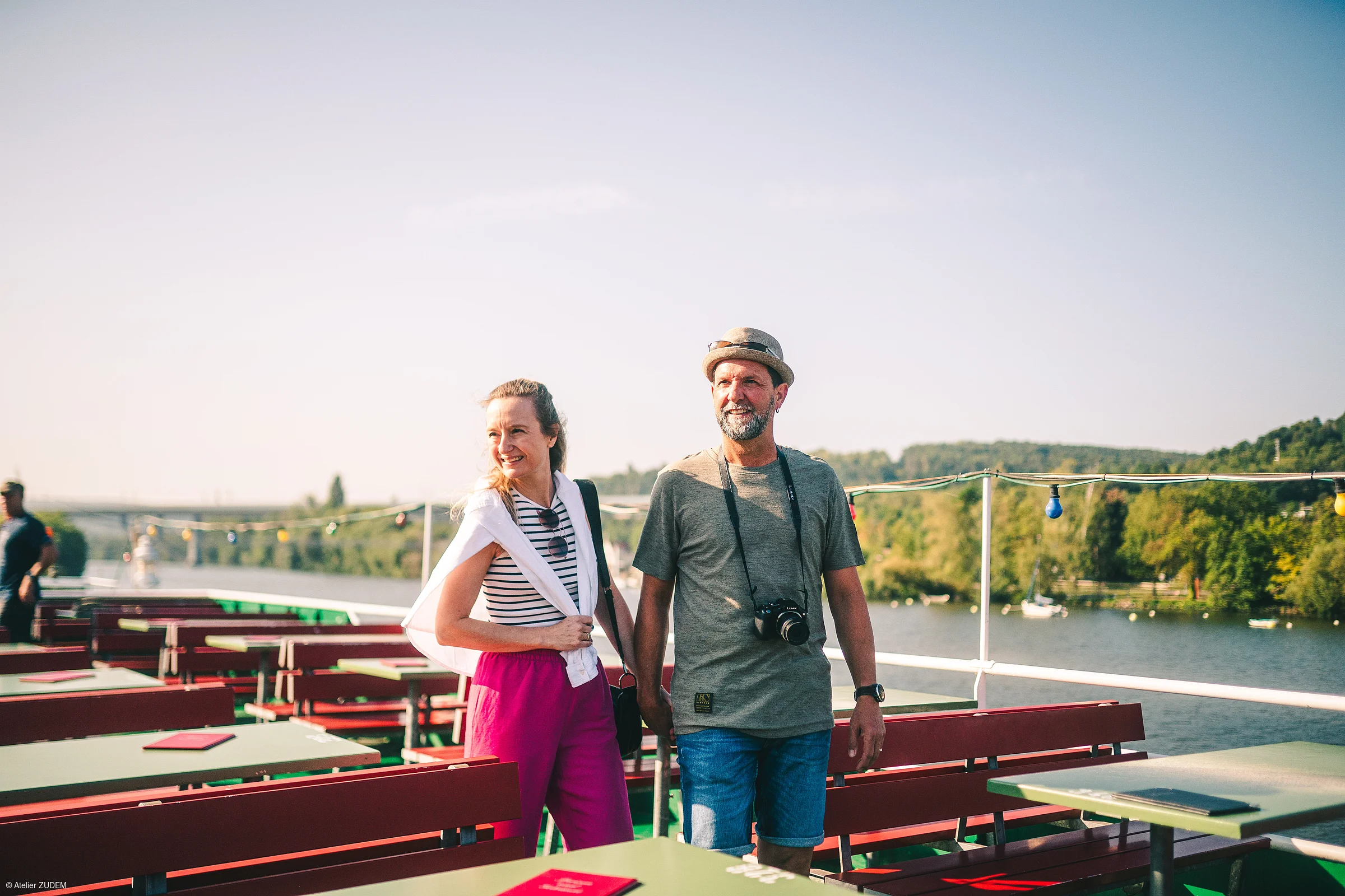 Paar geht auf Terrasse mit roten Bänken und grünen Tischen am Fluss bei Sonnenschein spazieren.