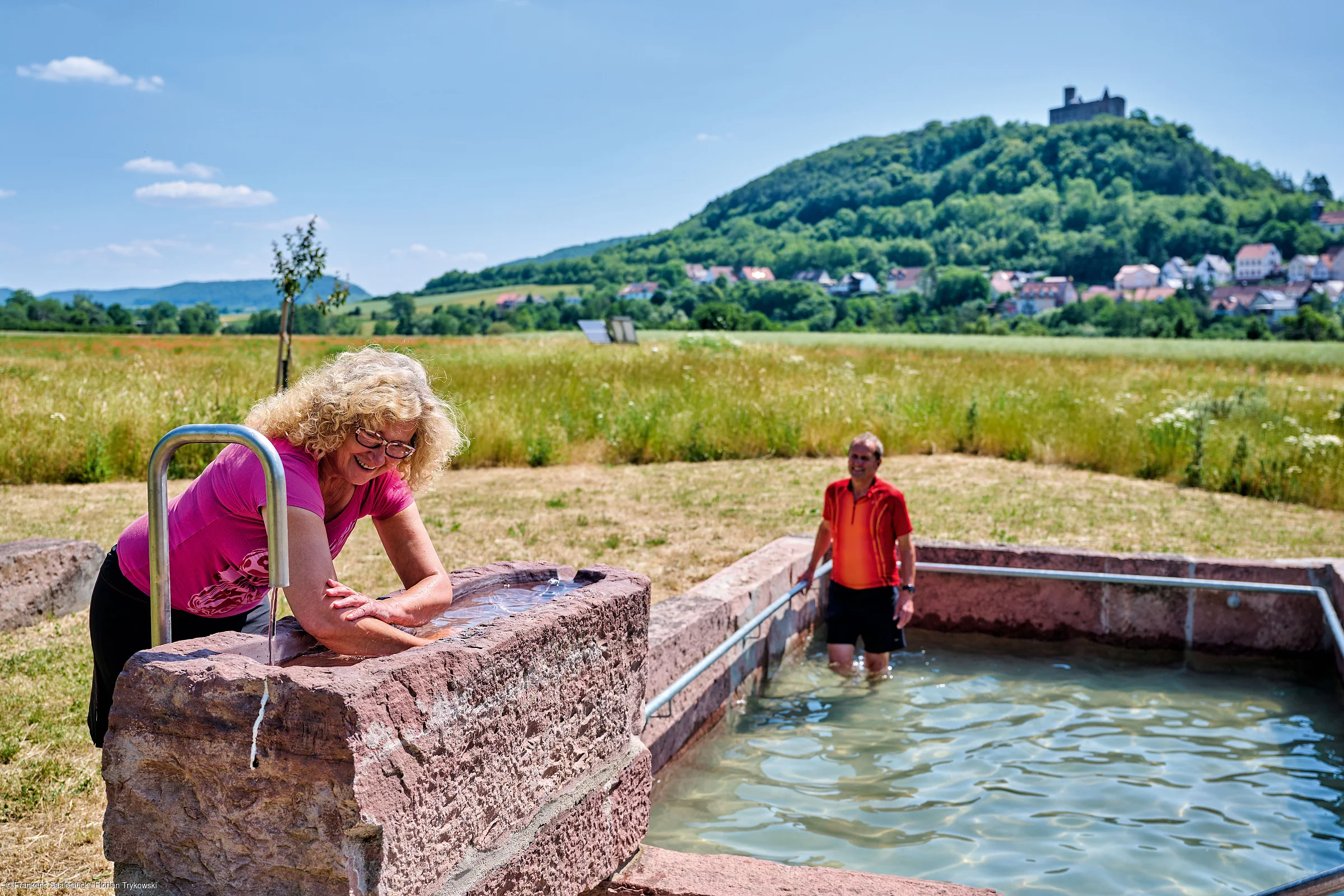Frau wäscht Arme an Brunnen, Mann steht im Wasserbecken, Wiese und bewaldeter Hügel mit Burg im Hintergrund.