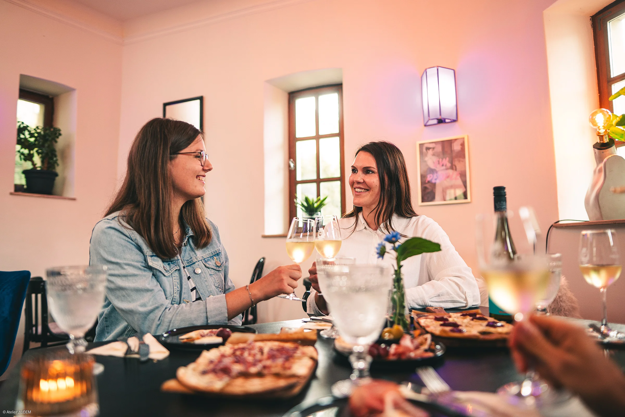 Zwei Frauen stoßen mit Weißweingläsern an einem gedeckten Tisch mit Essen und Getränken an.