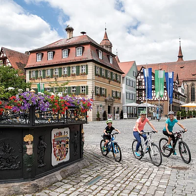 Drei Radfahrer fahren auf einem gepflasterten Platz mit historischen Gebäuden und einer Kirche im Hintergrund.