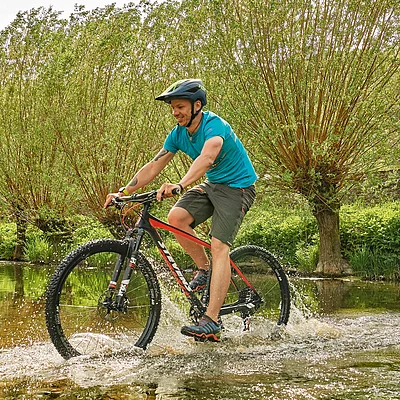 Mann mit Helm fährt Mountainbike durch flaches Wasser vor grünen Büschen und altem Gebäude.