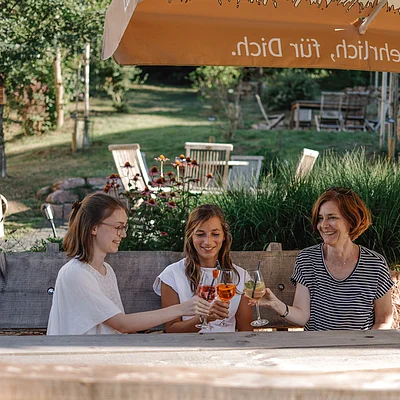 Drei Frauen sitzen draußen an einem Holztisch und stoßen mit Getränken unter einem Sonnenschirm an.