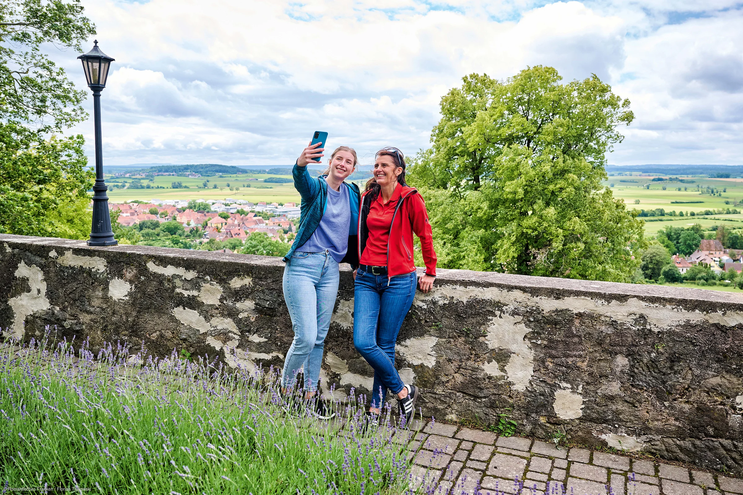 Zwei Frauen machen an einer Steinmauer mit Blick auf Landschaft und Dorf ein Selfie bei bewölktem Himmel.