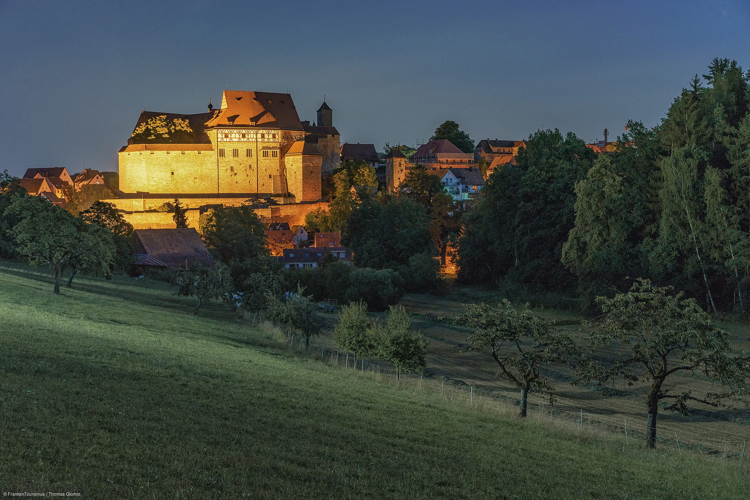 Beleuchtete Burg auf einem Hügel mit Bäumen und Wiesen im Vordergrund bei Abenddämmerung