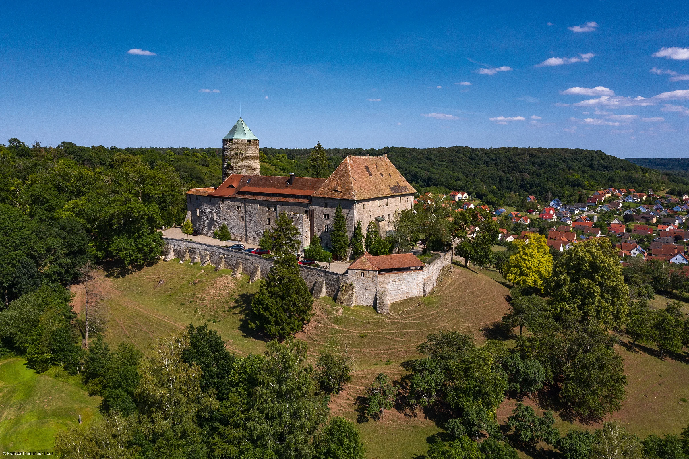 Luftaufnahme einer Burg auf einem bewaldeten Hügel mit Dorf im Hintergrund unter blauem Himmel.