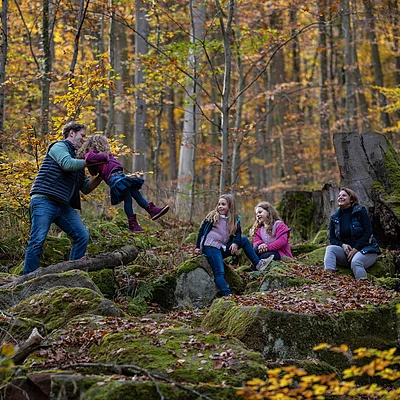 Vater spielt mit Kind, zwei Mädchen und Frau sitzen auf moosbedeckten Steinen im herbstlichen Wald.