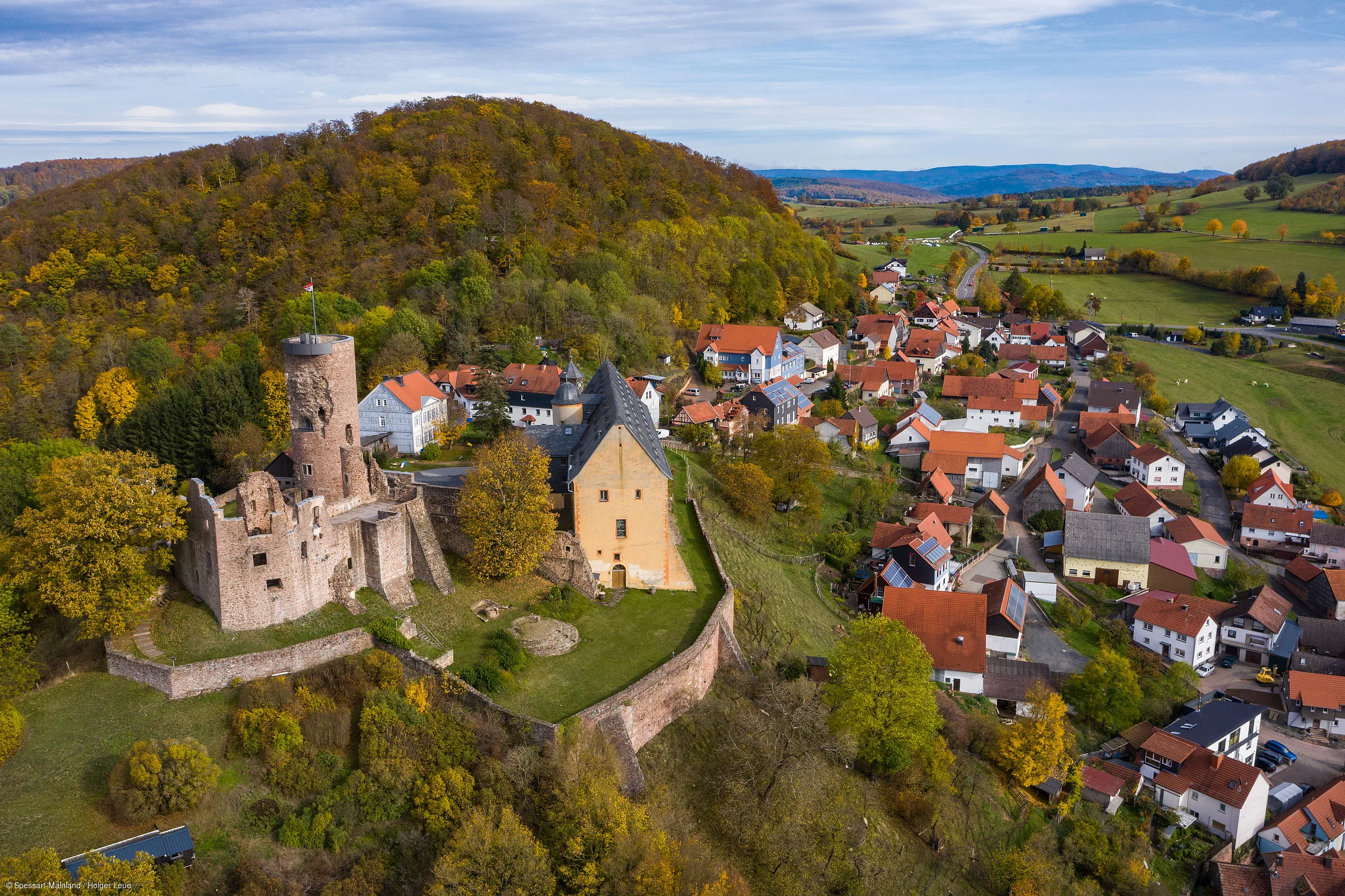 Luftaufnahme einer Burganlage mit umliegendem Dorf und herbstlich gefärbtem Wald auf einem Hügel.