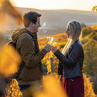 Zwei Personen stoßen mit Weingläsern in einem herbstlichen Weinberg bei Sonnenuntergang an.