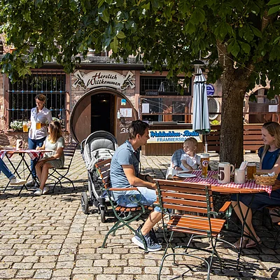 Außenbereich eines Cafés mit Gästen an Tischen auf gepflastertem Platz unter Baum und Sonnenschirm.