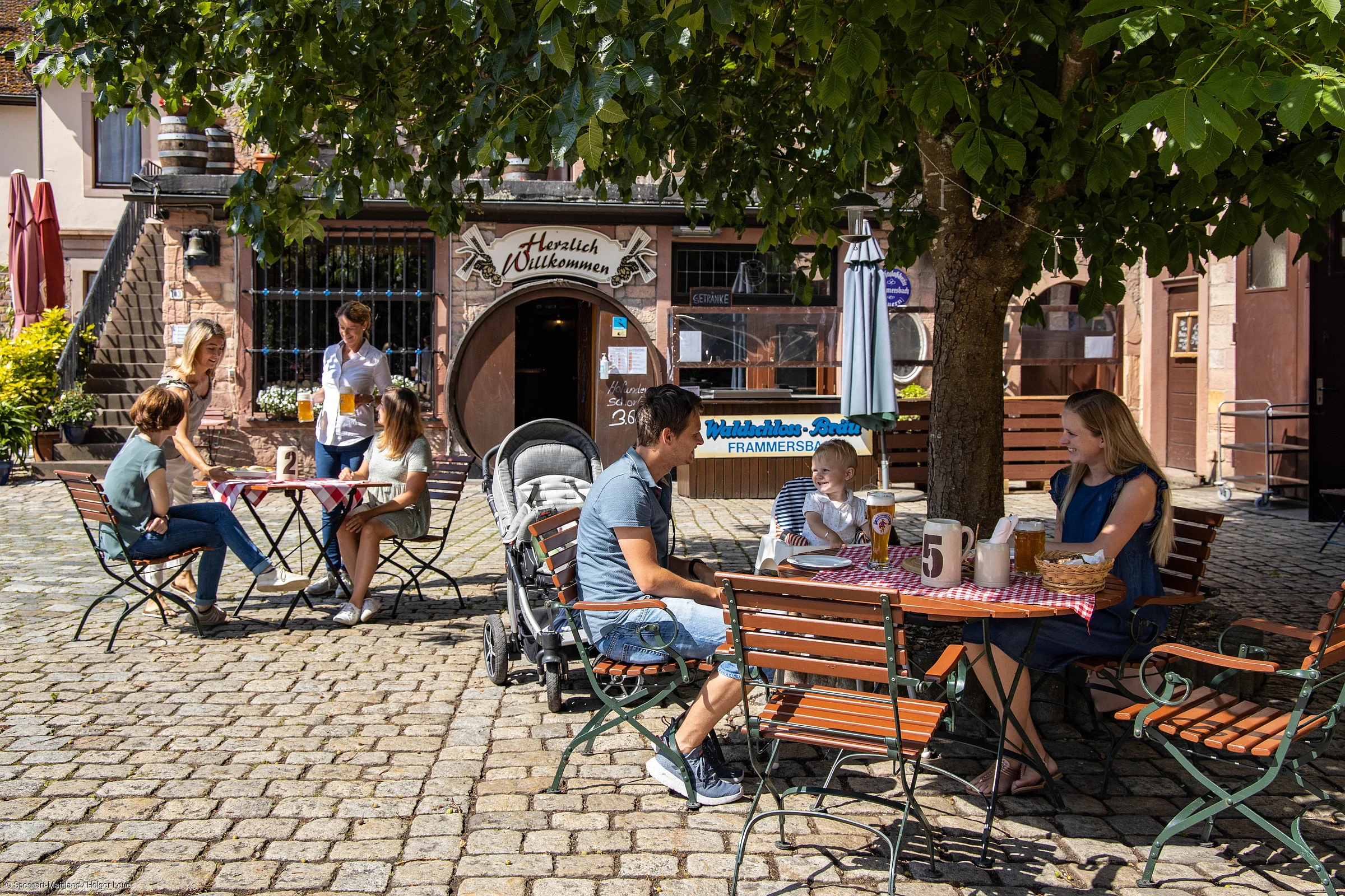Außenbereich eines Cafés mit Gästen an Tischen auf gepflastertem Platz unter Baum und Sonnenschirm.