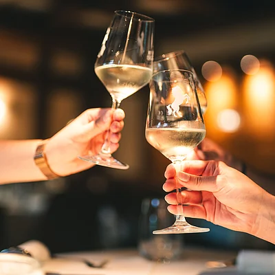 Drei Hände stoßen mit gefüllten Weingläsern in einem Restaurant an, unscharfer Hintergrund.