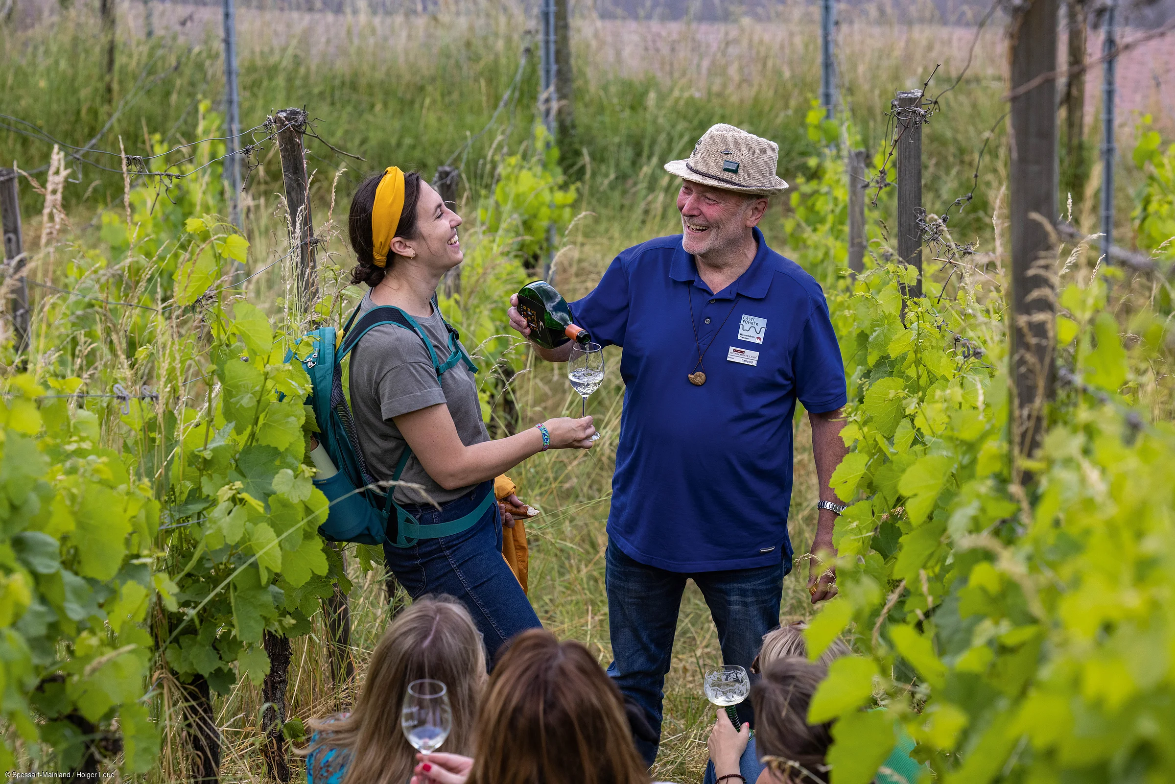 Mann schenkt Frau in Weinberg Weißwein in Glas ein, mehrere Personen beobachten die Szene.