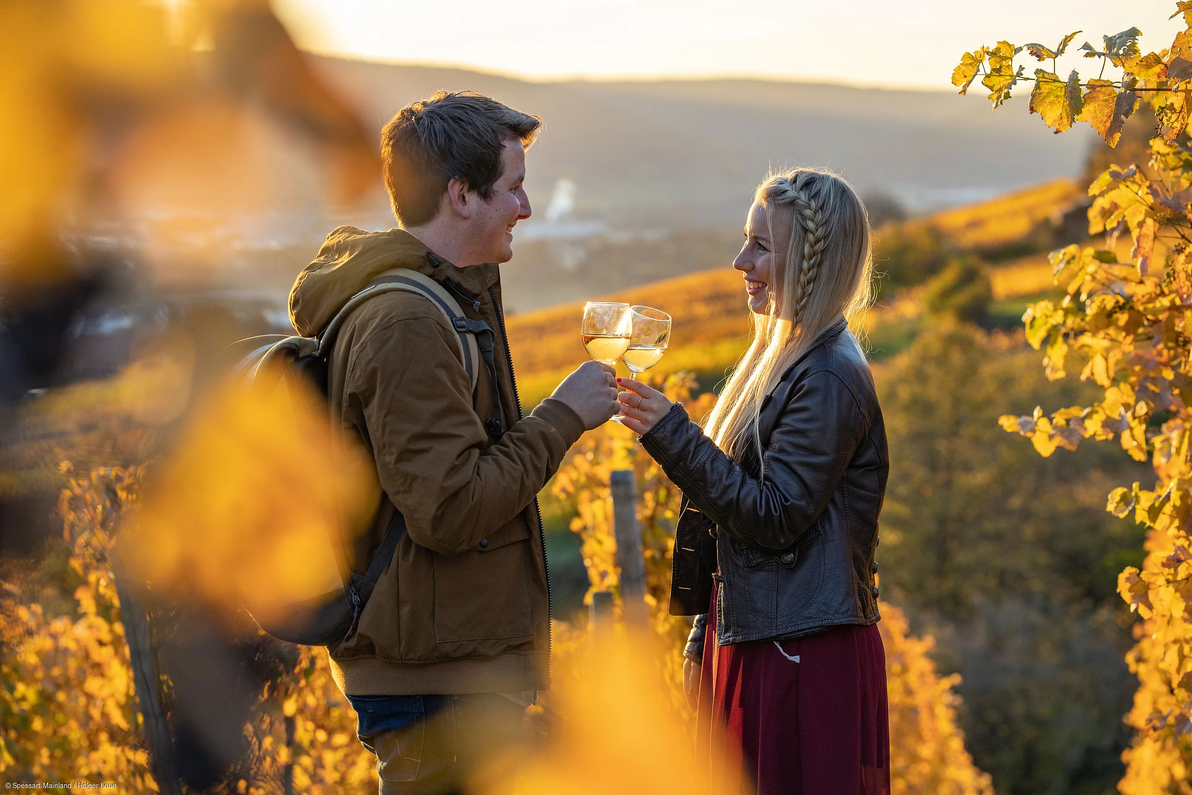 Zwei Personen stoßen mit Weingläsern in einem herbstlichen Weinberg bei Sonnenuntergang an.