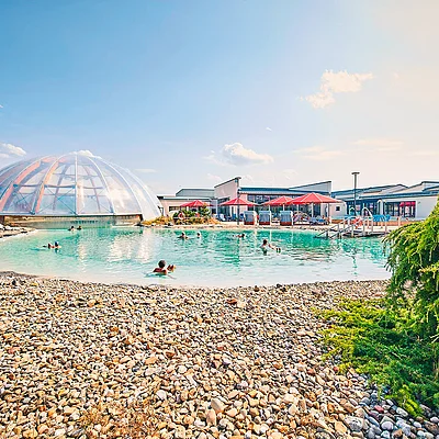 Außenpool mit Menschen, Kiesstrand, transparentes Kuppelgebäude und Gebäude mit Sonnenschirmen im Hintergrund.