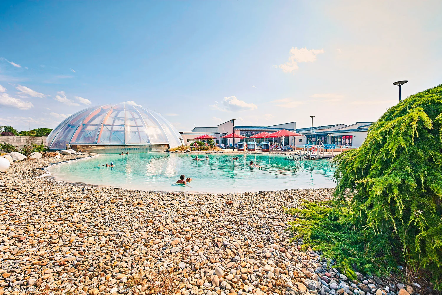 Außenpool mit Menschen, Kiesstrand, transparentes Kuppelgebäude und Gebäude mit Sonnenschirmen im Hintergrund.