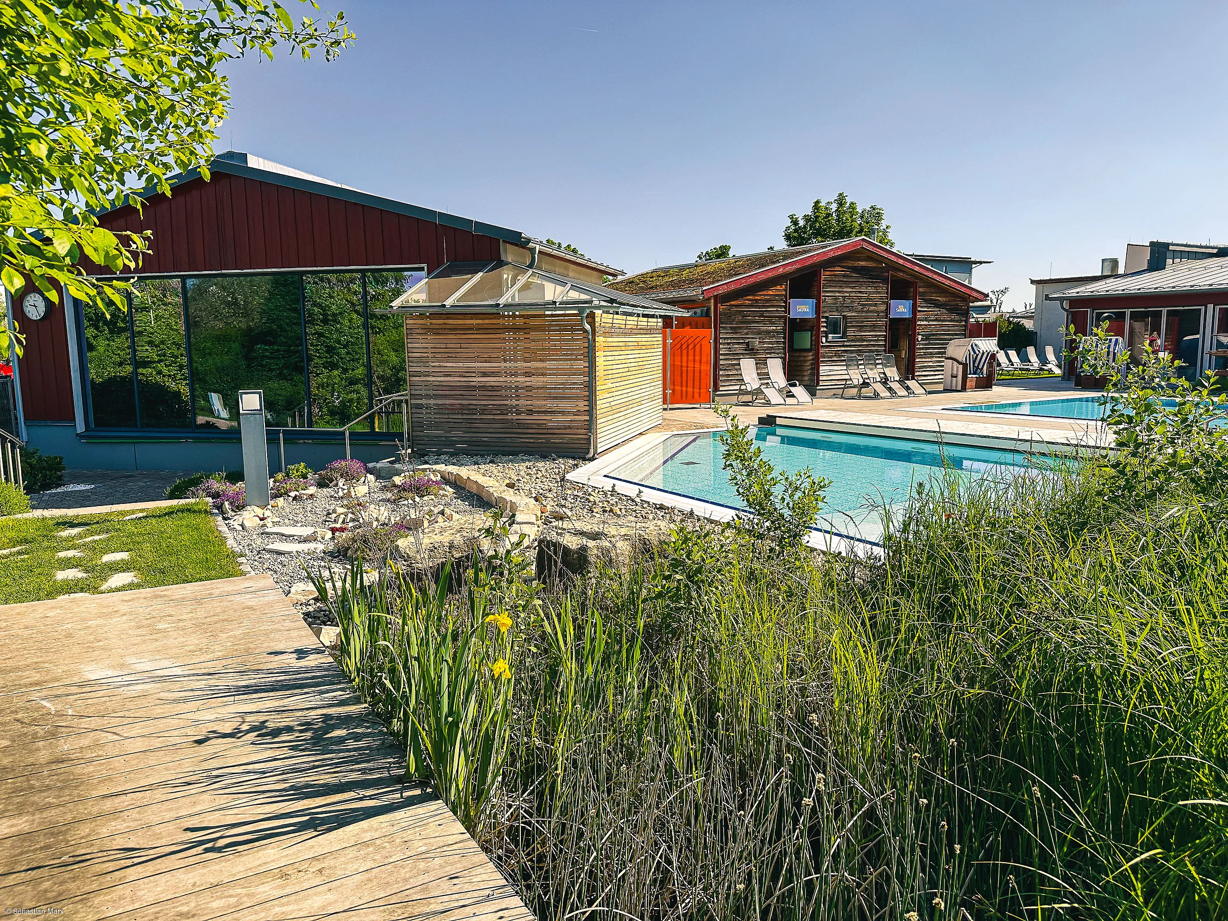 Außenbereich mit Schwimmbecken, Liegestühlen, Holzsteg und Gebäuden unter klarem Himmel.
