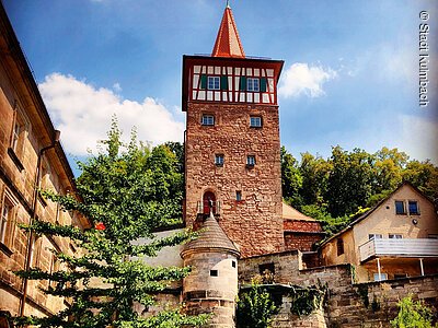 City view with Roter Turm (Kulmbach, Franconian Forest) Historischer Steinturm mit rotem Dach und Fachwerk, umgeben von Bäumen und weiteren Gebäuden bei blauem Himmel.